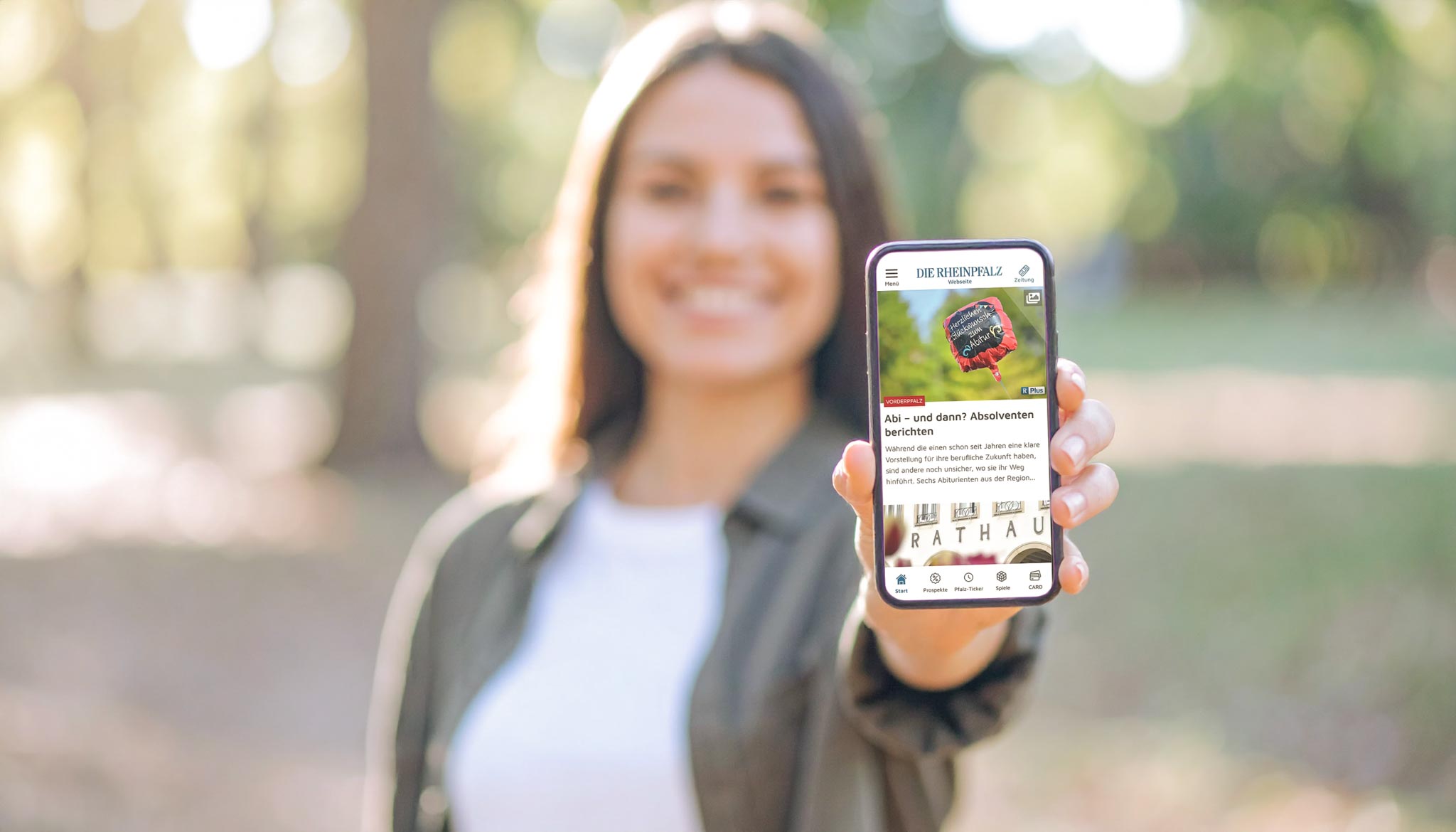 Junge Frau h&auml;lt l&auml;chelnd ein Smartphone mit der ge&ouml;ffneten RHEINPFALZ-App in die Kamera &ndash; Symbol f&uuml;r mobile Nutzung und digitalen Zugang