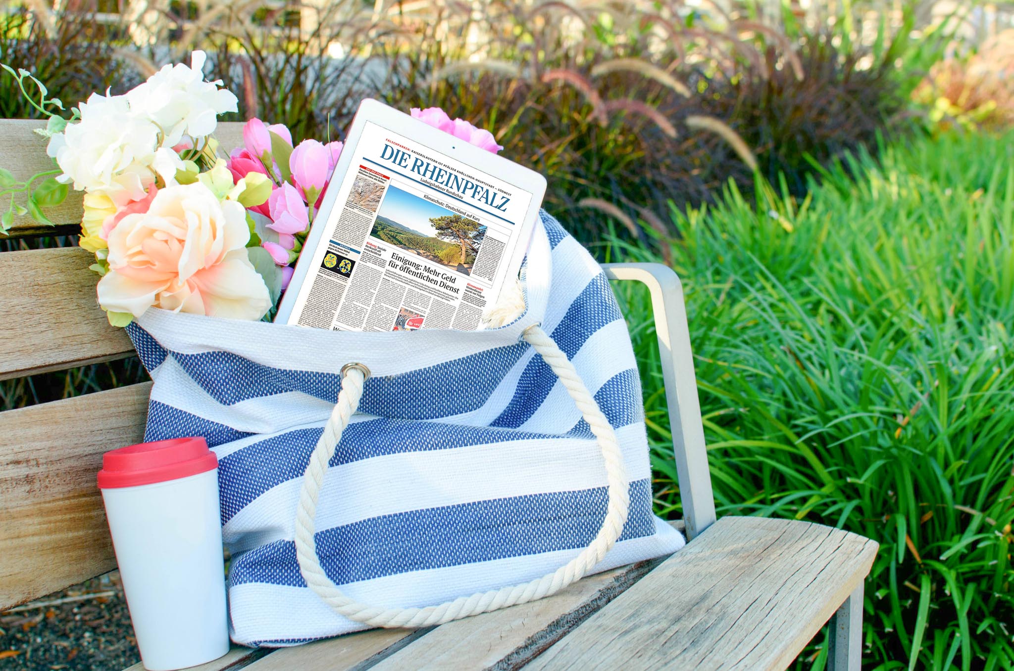 DIE RHEINPFALZ als Geschenk &ndash; Tablet mit der RHEINPFALZ in einer dekorativen Tasche mit Blumen. Daneben ein Thermobecher.